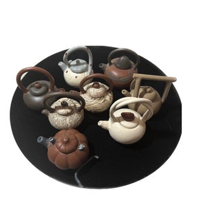 May include: A collection of eight small ceramic teapots in various colors and patterns, displayed on a black surface.