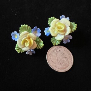Op de afbeelding: Een paar vintage bloemenoorbellen met gele rozen en blauwe bloemen. De oorbellen zijn gemaakt van porselein en hebben een delicaat, vrouwelijk ontwerp.