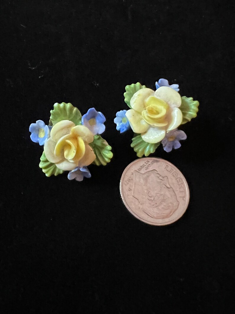 Op de afbeelding: Een paar vintage bloemenoorbellen met gele rozen en blauwe bloemen. De oorbellen zijn gemaakt van porselein en hebben een delicaat, vrouwelijk ontwerp.