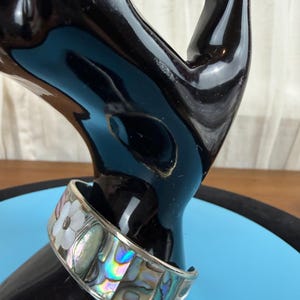 May include: A silver cuff bracelet with a floral design and iridescent abalone shell inlays. The bracelet features a white flower and rectangular shell pieces. The bracelet is displayed on a black hand-shaped stand against a blue background.