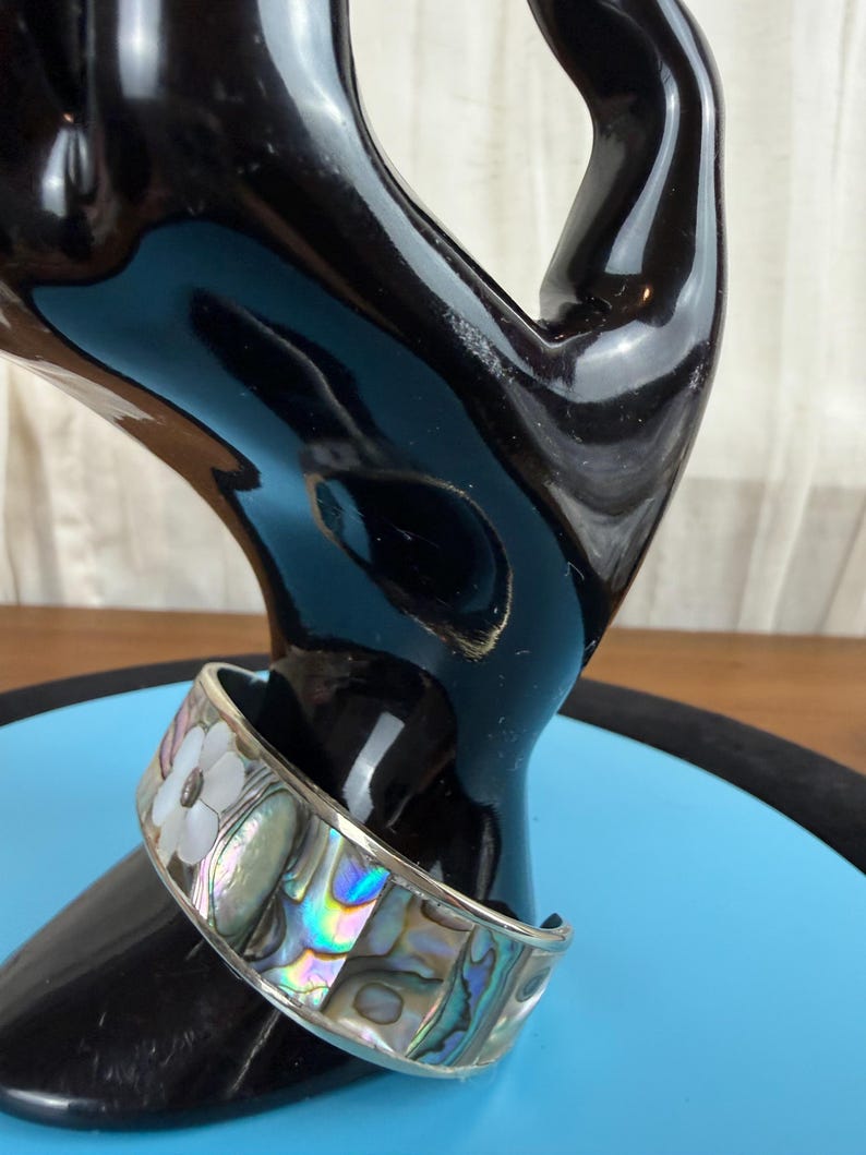 May include: A silver cuff bracelet with a floral design and iridescent abalone shell inlays. The bracelet features a white flower and rectangular shell pieces. The bracelet is displayed on a black hand-shaped stand against a blue background.