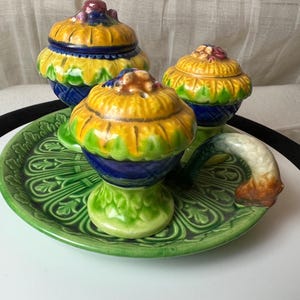 May include: A set of three ceramic salt and pepper shakers with a sunflower design. The shakers are green, blue, and yellow with a brown handle. The shakers are on a green and white ceramic tray with a floral design.