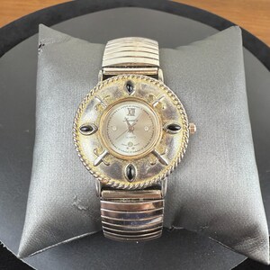 Op de afbeelding: Een zilverkleurig quartz horloge met een ronde wijzerplaat en een decoratieve goudkleurige lunette. Het horloge heeft zwarte accenten en een zilveren band. De wijzerplaat toont Romeinse cijfers en het woord "Quartz".