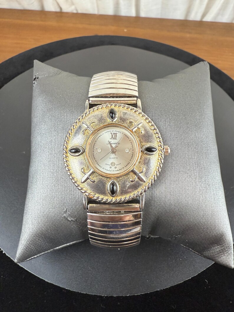 Op de afbeelding: Een zilverkleurig quartz horloge met een ronde wijzerplaat en een decoratieve goudkleurige lunette. Het horloge heeft zwarte accenten en een zilveren band. De wijzerplaat toont Romeinse cijfers en het woord "Quartz".