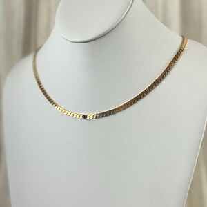 May include: A gold-colored herringbone chain necklace with a small dark blue stone. The necklace is displayed on a white mannequin bust against a neutral background. The chain is flat and has a subtle shine.