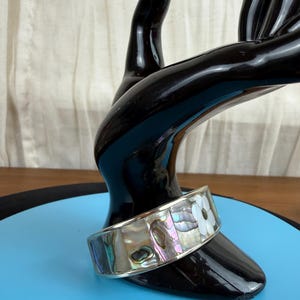 May include: A silver-toned cuff bracelet with iridescent abalone shell and white flower accents. The bracelet is displayed on a black hand-shaped stand against a blue background. The cuff is approximately 1.5 inches wide.
