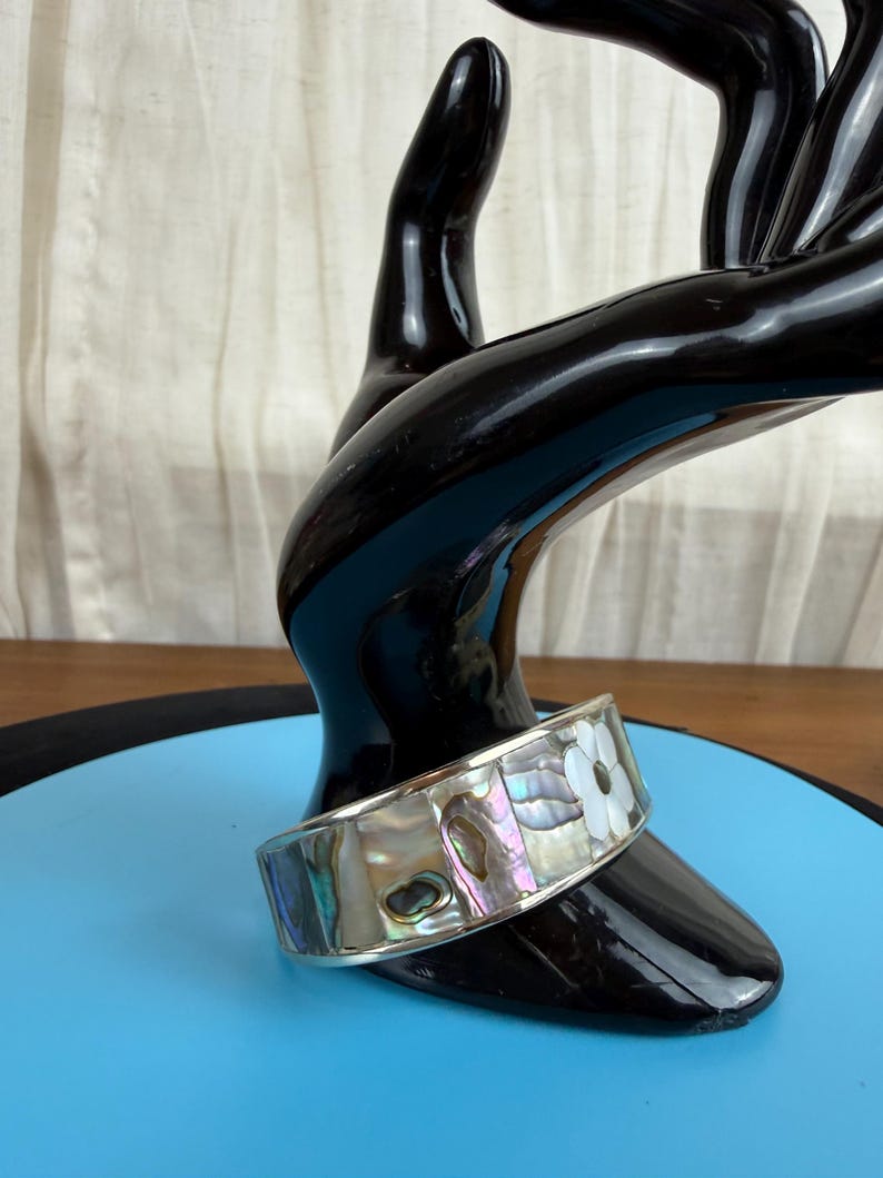 May include: A silver-toned cuff bracelet with iridescent abalone shell and white flower accents. The bracelet is displayed on a black hand-shaped stand against a blue background. The cuff is approximately 1.5 inches wide.
