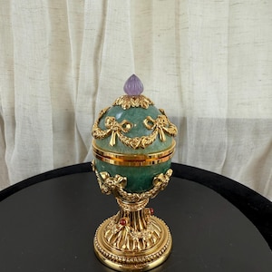 May include: An ornate, egg-shaped decorative object with a green, textured surface and gold accents. The egg features a gold base and a purple, teardrop-shaped top. Red jewels are set into the base. The object is likely a decorative item or a jewelry box.