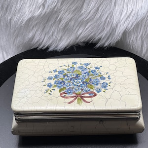 May include: A white ceramic trinket box with a floral design. The box has a cracked glaze finish and a silver trim around the edges. The floral design features a bouquet of blue forget-me-not flowers with a red ribbon.