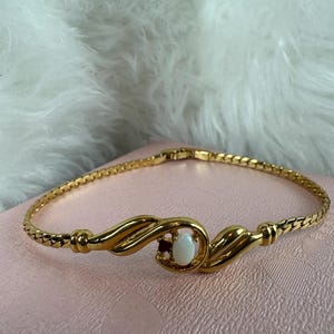 May include: A gold-toned bracelet with a central oval-shaped white stone. The bracelet features a twisted chain design and decorative gold swirls around the stone. The bracelet is displayed on a pink surface.
