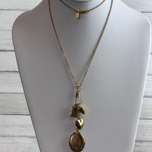 May include: A gold-tone necklace featuring a long chain with a multi-charm pendant. The pendant includes a small rectangular charm, a heart-shaped charm, and a larger oval locket with an embossed floral-style border design on the front surface.