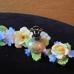 Op de afbeelding: Een paar vintage bloemenoorbellen met blauwe en witte bloemen en gele centra. De oorbellen zijn gemaakt van porselein en hebben een zilverkleurige metalen sluiting. De achterkant van de oorbellen is gemarkeerd met "Made in England".