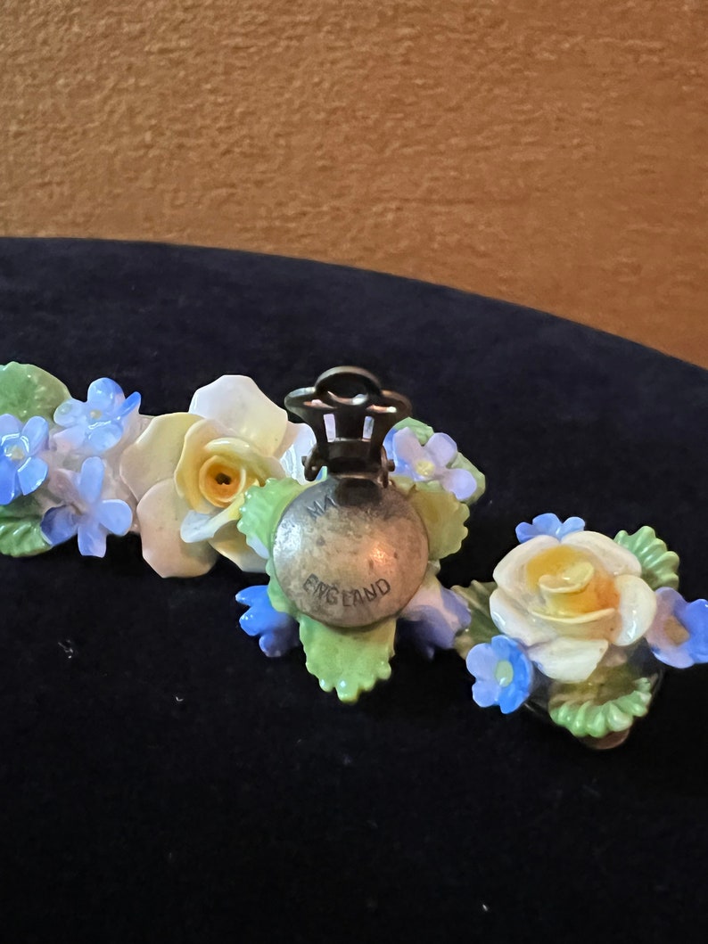 Op de afbeelding: Een paar vintage bloemenoorbellen met blauwe en witte bloemen en gele centra. De oorbellen zijn gemaakt van porselein en hebben een zilverkleurige metalen sluiting. De achterkant van de oorbellen is gemarkeerd met "Made in England".