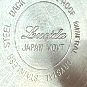 Op de afbeelding: Close-up van een zilveren horlogekast met de merknaam "Lucida" in cursief. De tekst "JAPAN MOVT", "STAINLESS STEEL BACK", "SHOWERPROOF" en "MINERAL CRYSTAL" zijn ook zichtbaar.