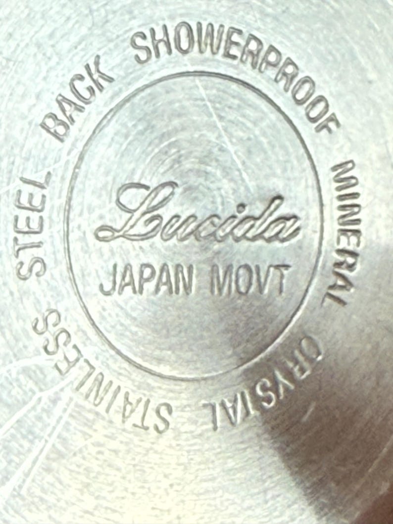 Op de afbeelding: Close-up van een zilveren horlogekast met de merknaam "Lucida" in cursief. De tekst "JAPAN MOVT", "STAINLESS STEEL BACK", "SHOWERPROOF" en "MINERAL CRYSTAL" zijn ook zichtbaar.