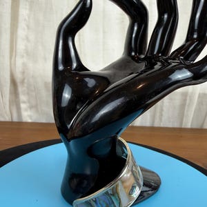 May include: A silver-toned cuff bracelet with an iridescent abalone shell inlay. The bracelet is displayed on a black hand-shaped sculpture against a blue surface. The abalone shell features a mix of blues, greens, and purples.
