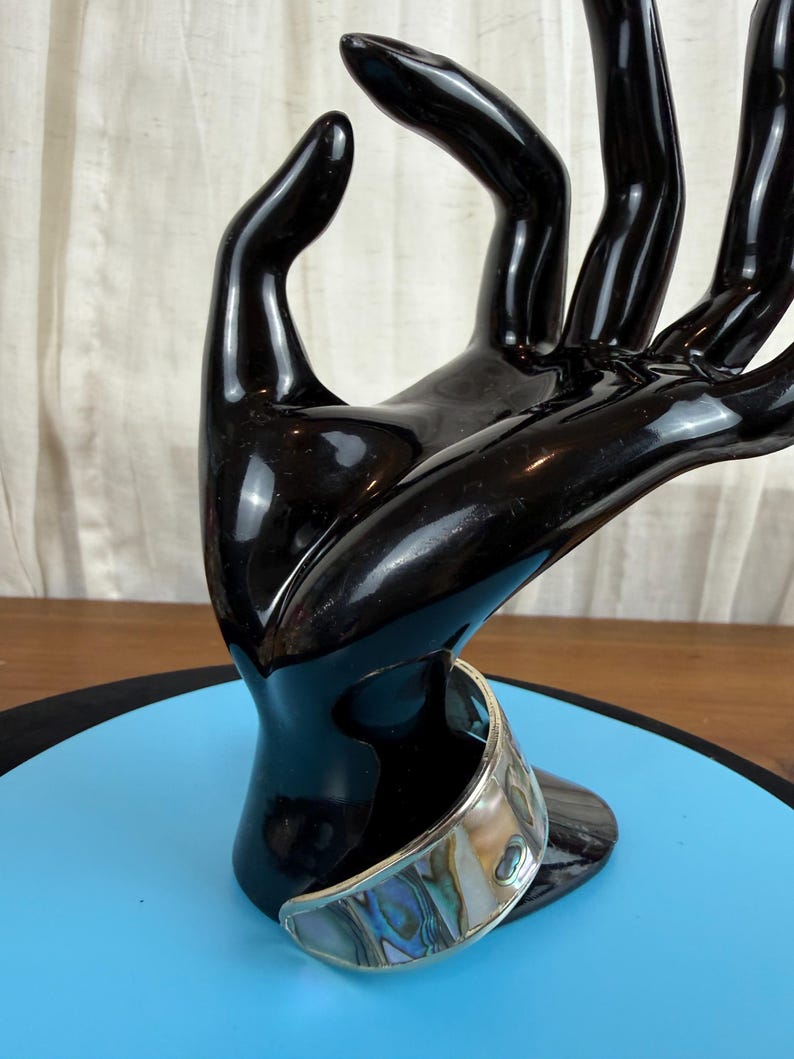 May include: A silver-toned cuff bracelet with an iridescent abalone shell inlay. The bracelet is displayed on a black hand-shaped sculpture against a blue surface. The abalone shell features a mix of blues, greens, and purples.