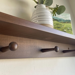 May include: A dark brown wooden shelf with three pegs below. A vase with a white and gray striped pattern and a small painting of a landscape are on the shelf. The shelf is mounted on a white wall.