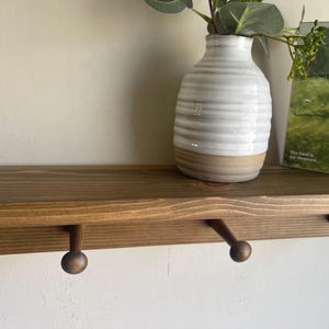 K&ouml;nnte beinhalten: Ein Holzregal mit drei runden Haken, einer wei&szlig;-beigen Vase mit Gr&uuml;nzeug und einem kleinen gerahmten Landschaftsbild. Das Regal ist in einem warmen Braunton gebeizt und an einer wei&szlig;en Wand befestigt.
