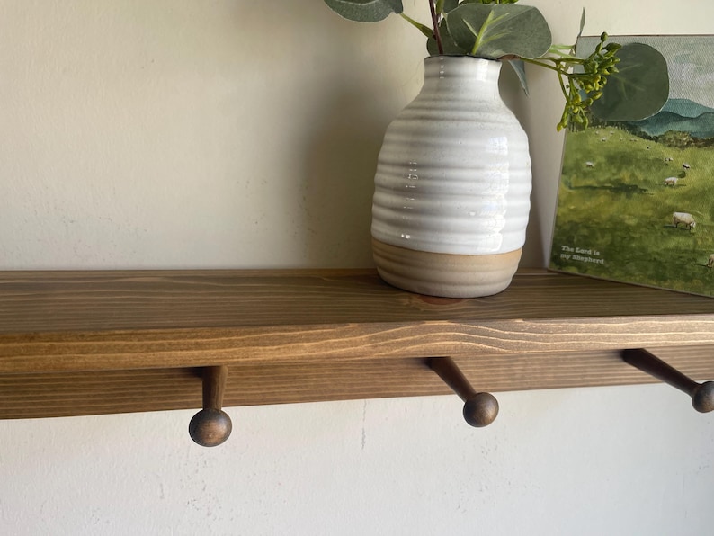 K&ouml;nnte beinhalten: Ein Holzregal mit drei runden Haken, einer wei&szlig;-beigen Vase mit Gr&uuml;nzeug und einem kleinen gerahmten Landschaftsbild. Das Regal ist in einem warmen Braunton gebeizt und an einer wei&szlig;en Wand befestigt.