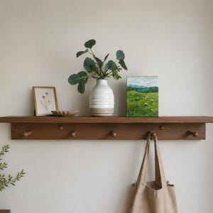 May include: A horizontal wooden wall shelf featuring six rounded pegs for hanging items. The shelf surface holds a white ribbed vase with green foliage, a small framed botanical print, a shallow scalloped dish, and a small landscape painting.