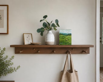 May include: A horizontal wooden wall shelf featuring six rounded pegs for hanging items. The shelf surface holds a white ribbed vase with green foliage, a small framed botanical print, a shallow scalloped dish, and a small landscape painting.
