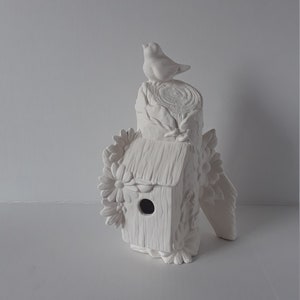 May include: White ceramic birdhouse with a bird perched on top, surrounded by flowers. The birdhouse is on a tree stump.