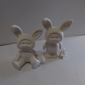 May include: Two white ceramic bunny figurines, each wearing a bunny costume with long ears. The figurines are sitting on a white surface.