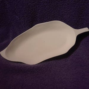 May include: A white, leaf-shaped ceramic dish with a handle. The dish has a smooth, matte finish and is set against a purple background. The dish is designed with a shallow bowl and a raised edge.