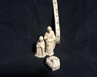 May include: White porcelain nativity scene figurines of Mary, Joseph, and baby Jesus in a manger. The figures are standing on a dark surface.
