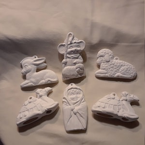 May include: A collection of unpainted ceramic Christmas ornaments. The set includes a donkey, a shepherd, a lamb, a camel, a baby wrapped in a blanket, and a bird. Each ornament has a hole for hanging.