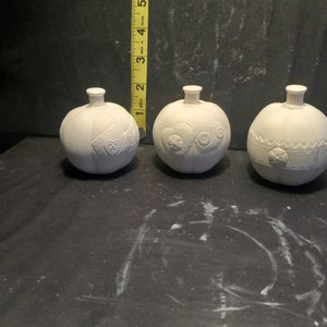May include: Three white ceramic bottle stoppers with different embossed designs. The stoppers are round and have a small knob on top.