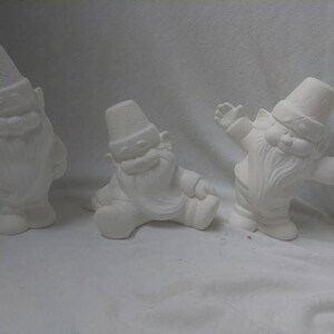 May include: Three white ceramic gnomes with different poses. The gnome on the left is standing with its arms crossed. The gnome in the middle is sitting with its legs crossed. The gnome on the right is standing with its arms outstretched.