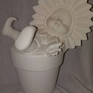 May include: A ceramic figurine of a baby in a sunflower costume, emerging from a white flower pot. The figurine is unpainted, with visible details of the baby's face, hands, and feet. The sunflower petals frame the baby's face.