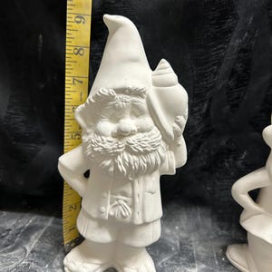 May include: A white ceramic gnome figurine, approximately 8 inches tall, holding a seashell. The gnome has a long white beard, a pointed hat, and is wearing a jacket and pants. The figurine is unpainted and ready for customization.
