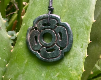 May include: A dark green, circular stone pendant featuring a carved, four-petaled floral design with a central hole. The pendant is suspended from a thin, black cord that passes through a small opening at the top of the stone piece.