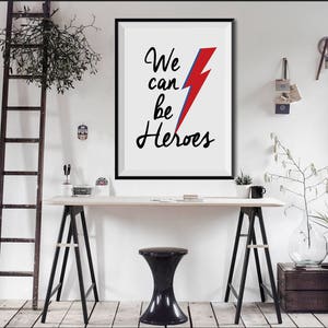 May include: A black framed print with a white background and the text "We can be Heroes" in black cursive writing. A red and blue lightning bolt is centered above the text.