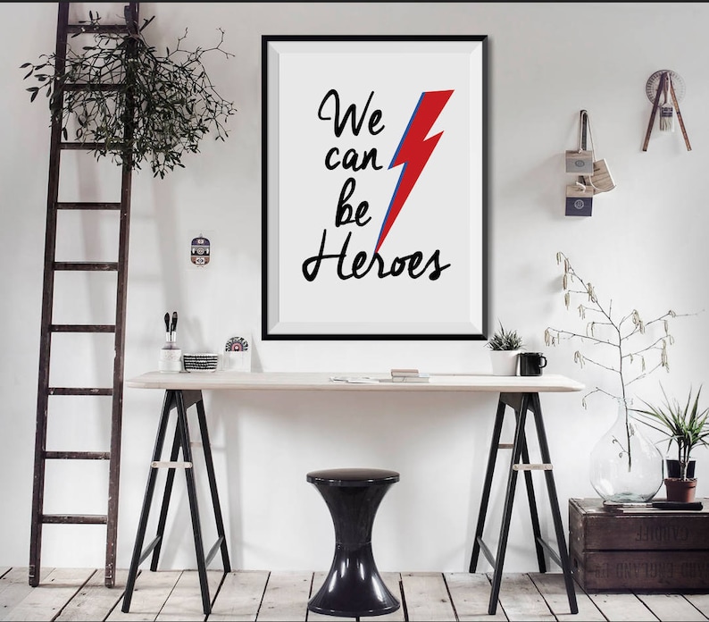 May include: A black framed print with a white background and the text "We can be Heroes" in black cursive writing. A red and blue lightning bolt is centered above the text.