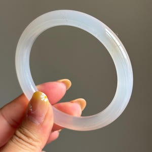May include: A smooth, translucent white bangle bracelet with a polished finish. The circular accessory features a consistent thickness and a rounded profile, showcasing a milky, semi-transparent appearance that allows light to pass through the material.
