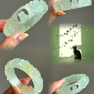 May include: A pale green translucent bangle bracelet featuring carved floral motifs and a small cat silhouette cutout. The smooth, polished surface displays intricate relief details of flowers encircling the band, creating a continuous pattern.