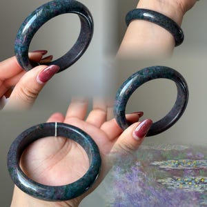 May include: A polished bangle bracelet crafted from a dark, mottled stone featuring deep blue, green, and magenta-pink inclusions. The circular band has a smooth, glossy finish and a rounded profile, showcasing natural color variations throughout.