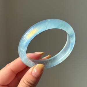 May include: A translucent, light blue bangle bracelet with a smooth, polished surface. The circular band features subtle internal variations and a soft, icy appearance that catches the light, creating a clear and luminous aesthetic.