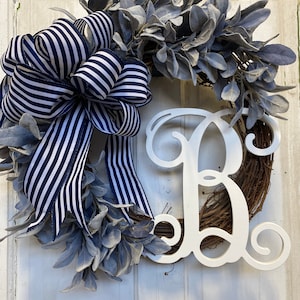 Puede incluir: Una corona hecha de hojas verde grisáceas con una letra "R" de madera blanca en el centro. La corona está decorada con un lazo de cinta a rayas blancas y negras.