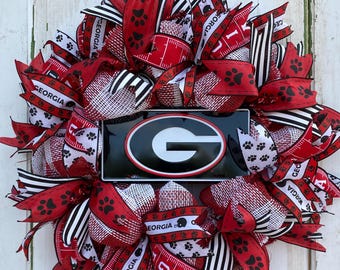 May include: A red, white, and black wreath with a black and white checkered ribbon, red ribbon with paw prints, and a red ribbon with the word "Georgia" on it. The wreath also has a black and white mesh and a black metal sign with a white "G" on it.