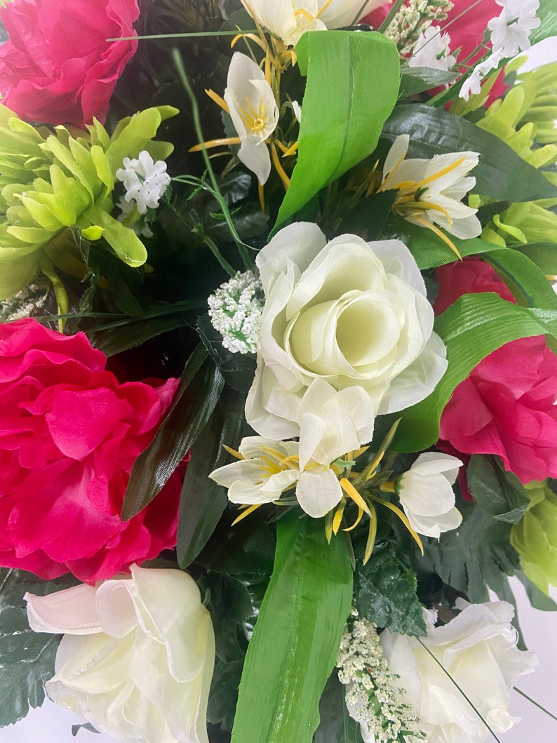 May include: A bouquet of artificial flowers with pink peonies, white roses, and small white flowers. The bouquet is surrounded by green leaves.