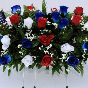 May include: A patriotic floral arrangement featuring red, white, and blue roses with green foliage and two American flags.