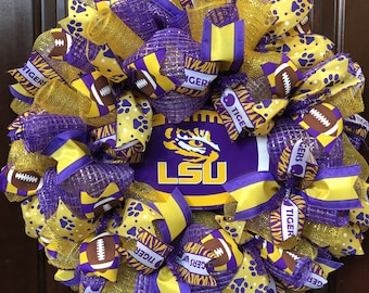 May include: A purple and gold wreath featuring an LSU logo in the centre. The wreath is decorated with purple and gold ribbons, footballs, and paw prints.
