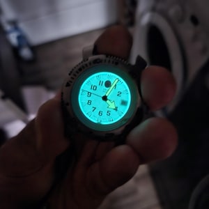 Könnte beinhalten: Nahaufnahme einer Armbanduhr mit leuchtend hellblauem Zifferblatt. Die Uhr hat eine silberfarbene Lünette und ein schwarzes Armband. Die Zeiger der Uhr zeigen die Uhrzeit an. Das Zifferblatt zeigt die Zahlen 1 bis 12.