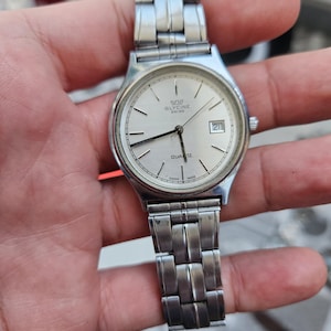 Può includere: Un orologio Glycine Swiss Quartz argentato con quadrante rotondo e datario. L'orologio ha un cinturino argentato e un quadrante argentato con lancette e indici neri. L'orologio è tenuto in una mano.