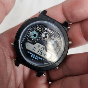 Könnte beinhalten: Schwarze digitale Casio-Uhr mit rundem Zifferblatt. Die Uhr zeigt die Uhrzeit 1:154 an und trägt den Text "WATER RESIST 200M". Die Uhr hat eine schwarze Lünette und eine Digitalanzeige.